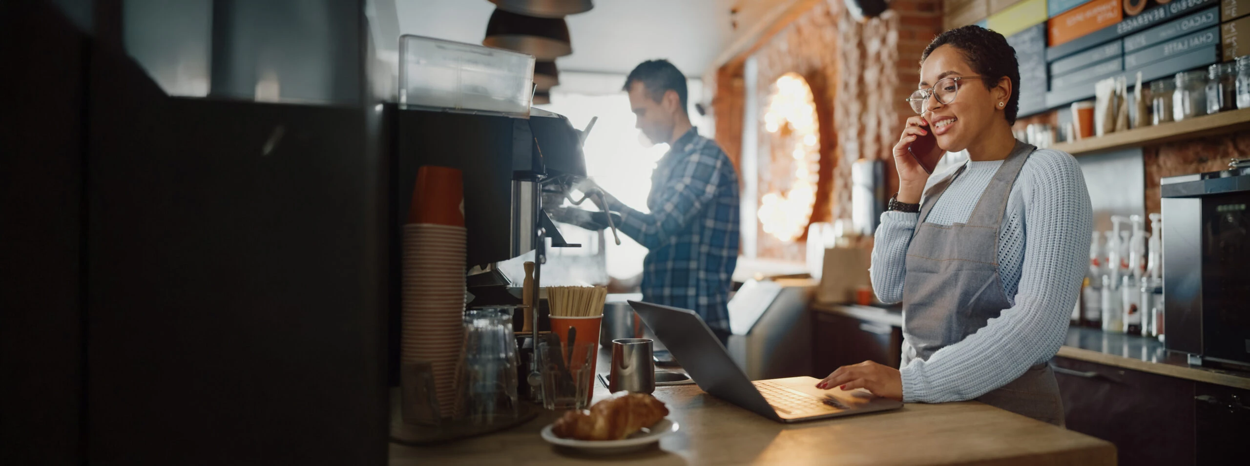 bistro employee talking on the phone while using a laptop and another employee makes a hot beverage in the background