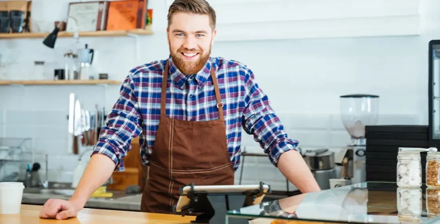 Barista in a checkered shirt and brown apron standing in a coffee shop next to a tablet POS system.