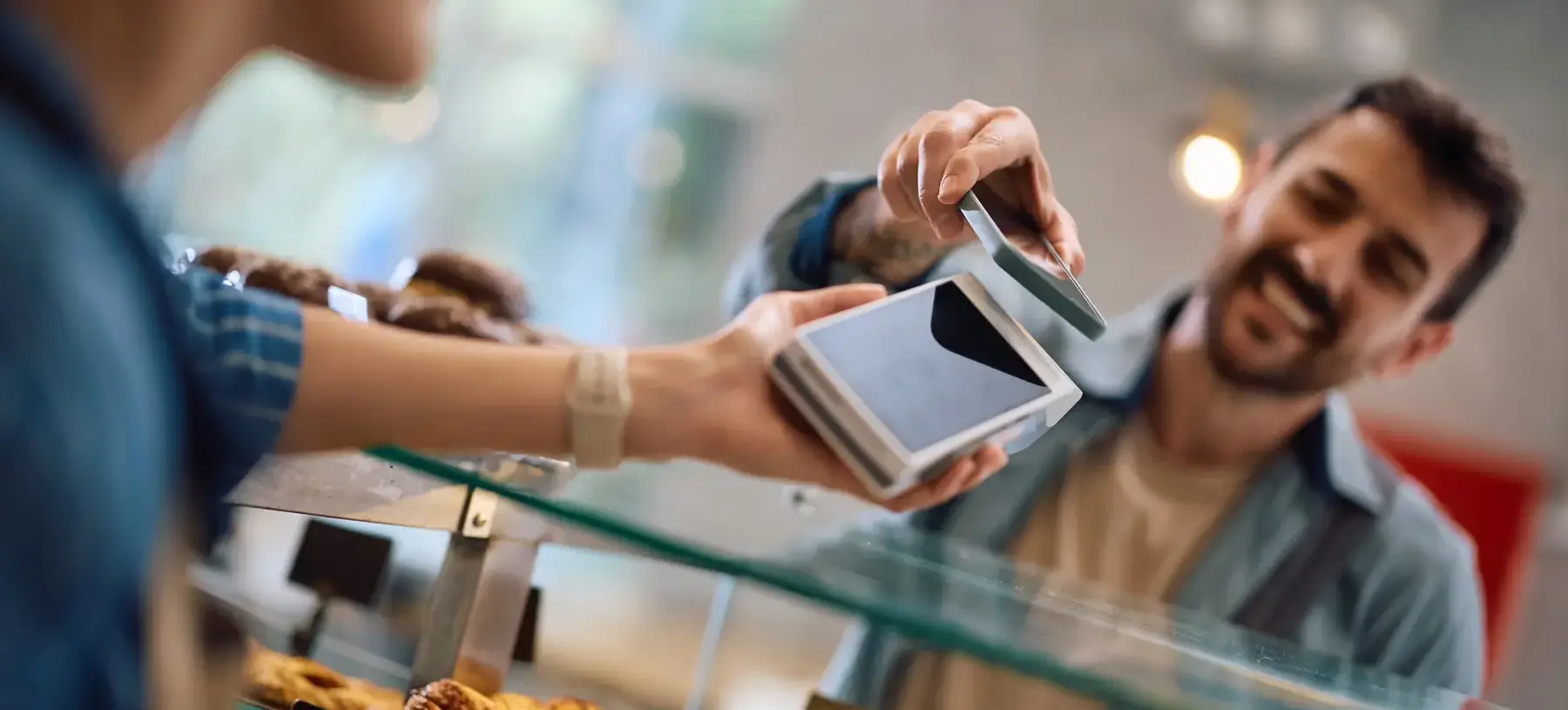 Cliente en una panadería pagando en el mostrador al acercar su teléfono a la terminal de pago sostenida por el empleado