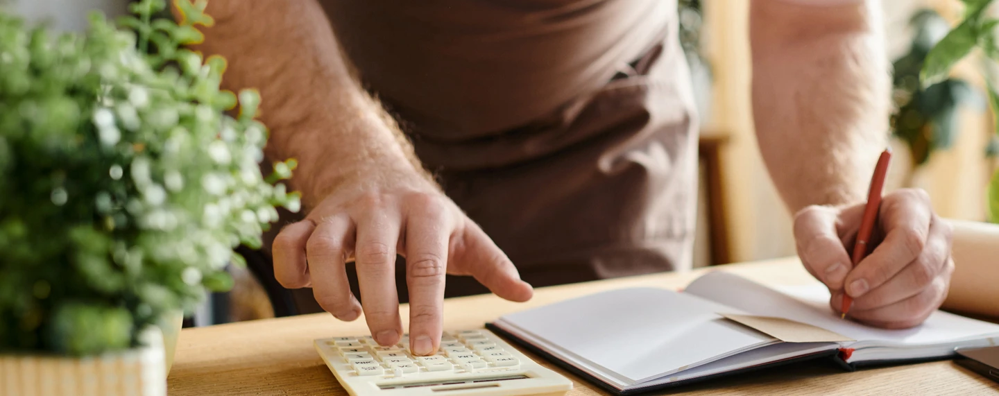 person standing at a desk writing in a notebook with their left hand and using a calculator with their right hand